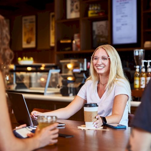 A smiling barista chats with customers at a cafe table, with laptops and coffee cups nearby.