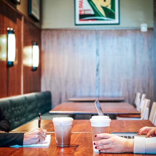 In a cozy caf&eacute;, people sit at a wooden table taking notes with coffee cups, with a framed poster on the wall behind them.