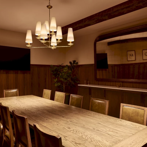 A modern dining room with a long wooden table, cushioned chairs, and glowing chandeliers, styled with warm wood walls and bar decor.