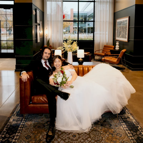 A newlywed couple poses in a modern hotel lounge, the bride in a white gown holding a bouquet and the groom holding champagne.