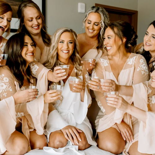 A group of bridesmaids in satin robes laugh and toast with champagne glasses together in a room.