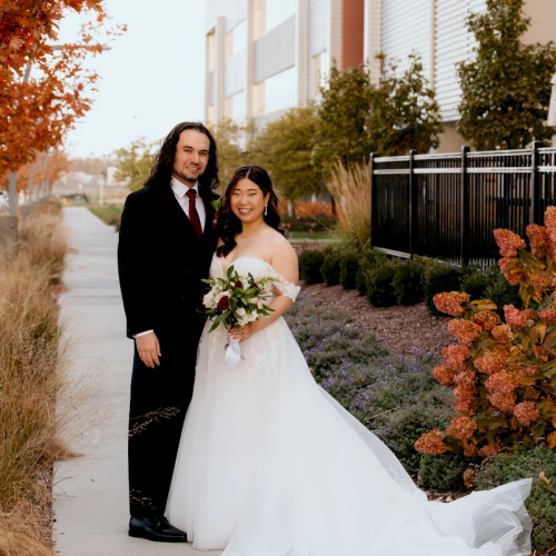 A bride and groom in formal wedding clothes stand outdoors on a walkway beside autumn plants and bushes.