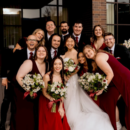 Wedding group photo: a bride in white with bouquets, surrounded by friends in red holding flowers.