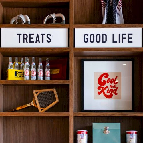 A wooden cabinet shelf displays signs &ldquo;TREATS&rdquo; and &ldquo;GOOD LIFE&rdquo; above drinks like Pepsi and a framed &ldquo;Eat to be Kind&rdquo; print.