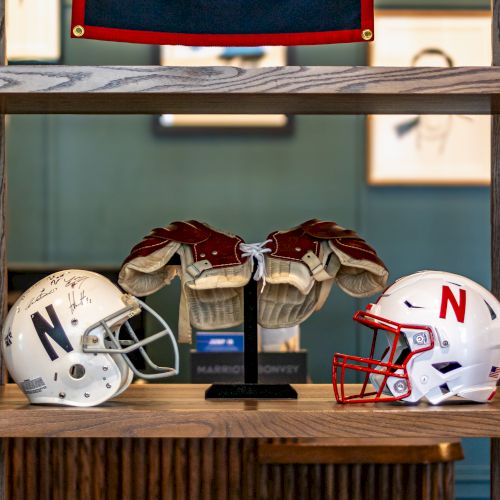Two football helmets sit on a wooden shelf, with a pair of brown-and-white shoulder pads and a red/blue banner above.