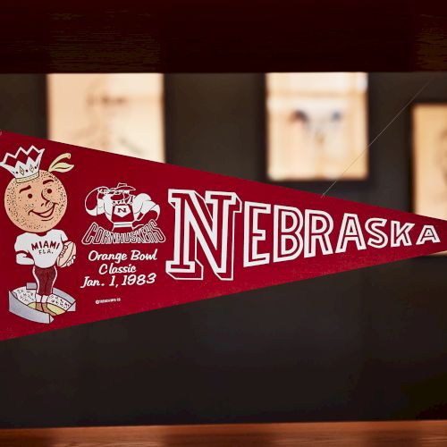 A framed red Nebraska Cornhuskers pennant for the Orange Bowl Classic Jan 1, 1983, with logos and a triangular banner.