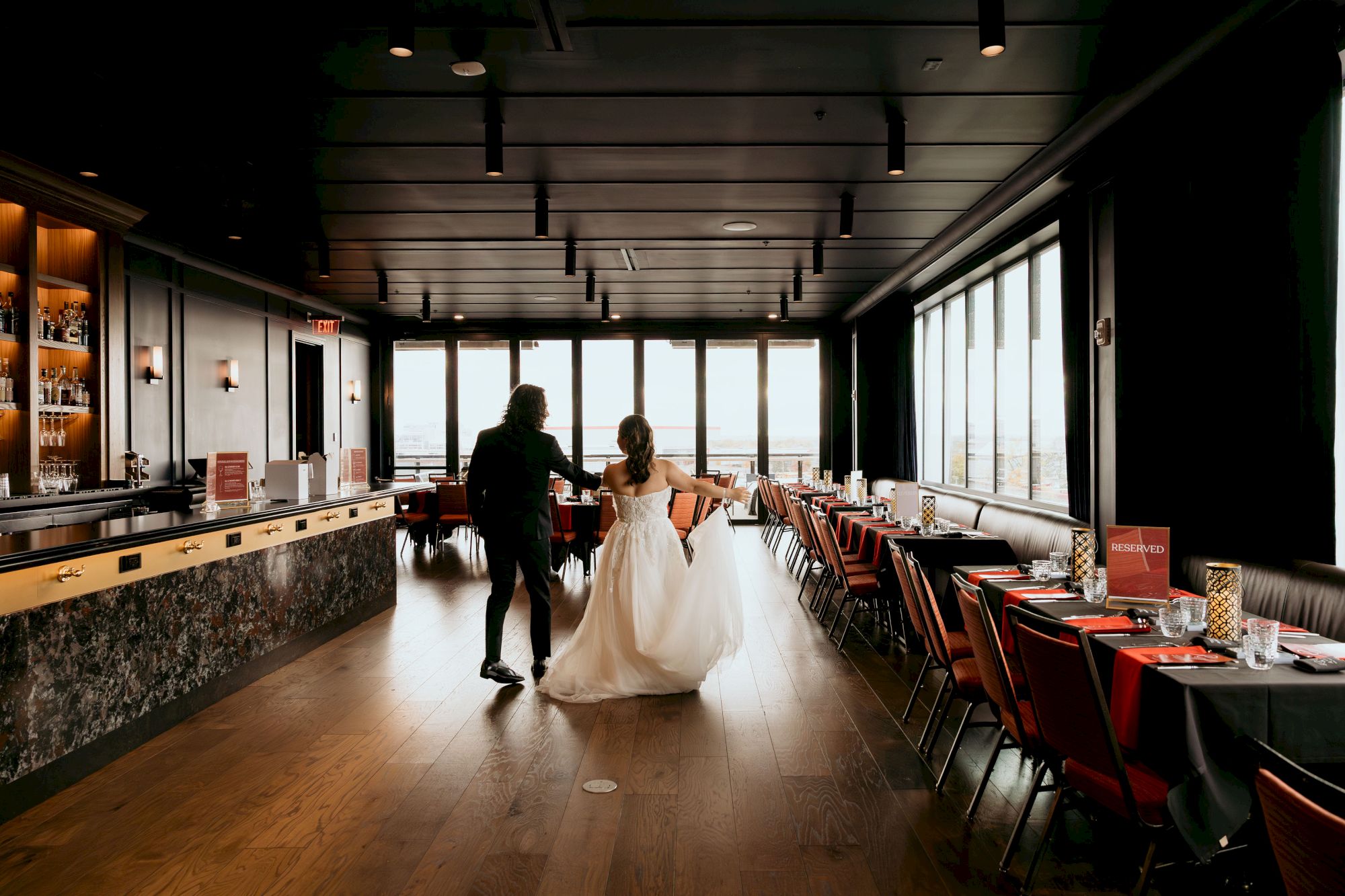 A couple in wedding attire walks hand-in-hand through a modern restaurant with a bar, tables set up, and big windows.