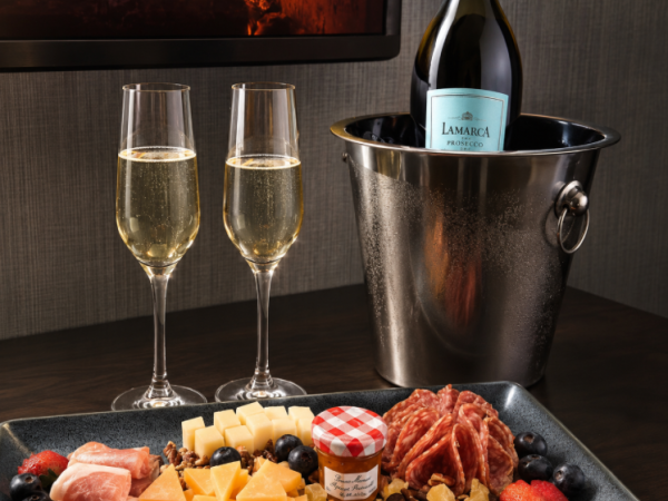 Two prosecco flutes and a Lamarka bottle sit beside a cheese/charcuterie tray with strawberries, grapes, and nuts by a fireplace.