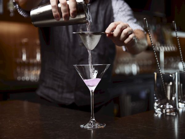 A bartender pours a purple cocktail into a martini glass with a strainer at a bar, with bottles in the background.
