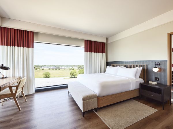 A bright hotel bedroom with a large bed, red-and-white curtains, and a window view of green fields.